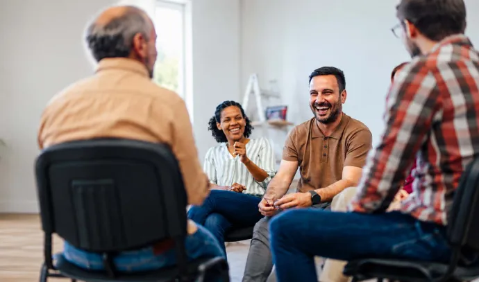 A group of adults sit in a circle during a supportive group discussion, smiling and engaging with one another.