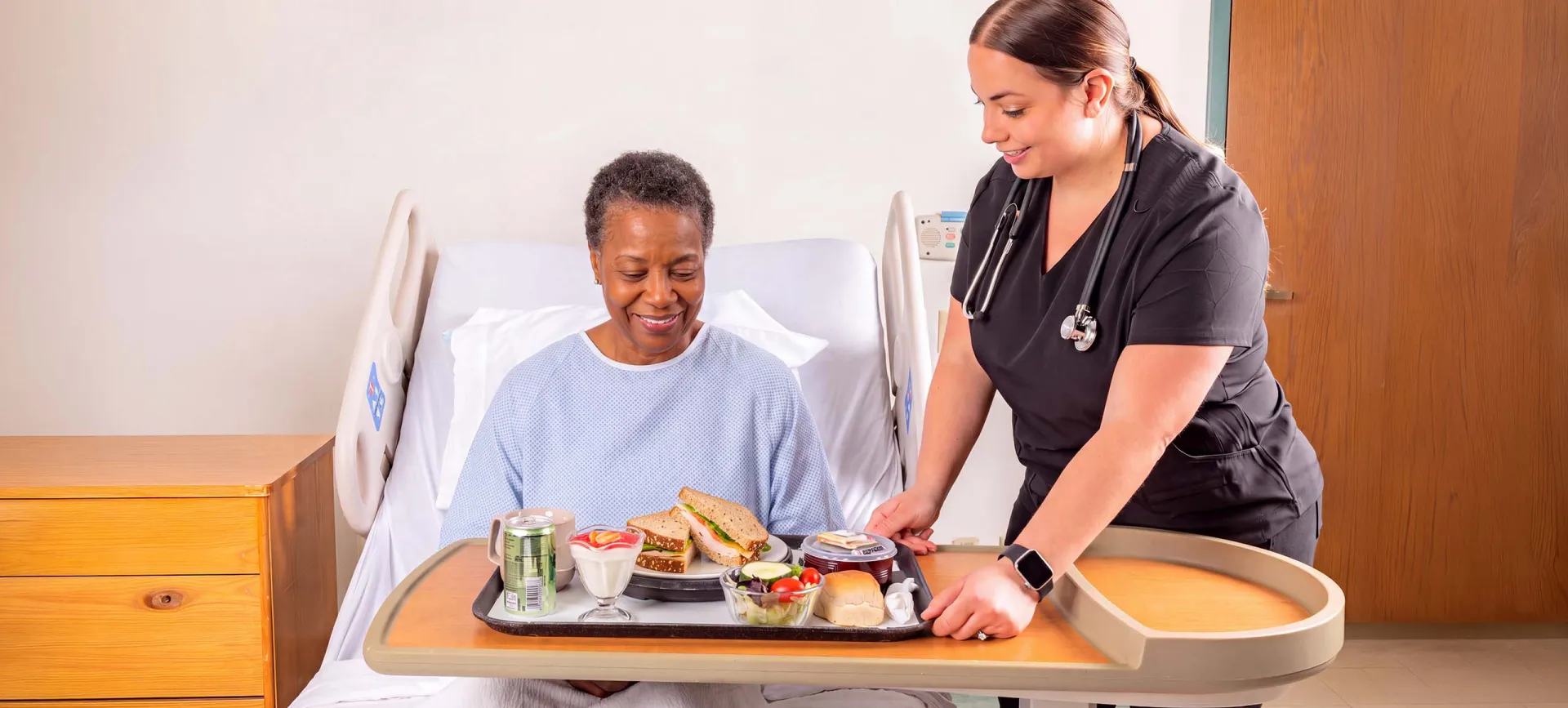 A woman in a hospital bed looks at a tray of food being served by a healthcare worker in a clinical setting.