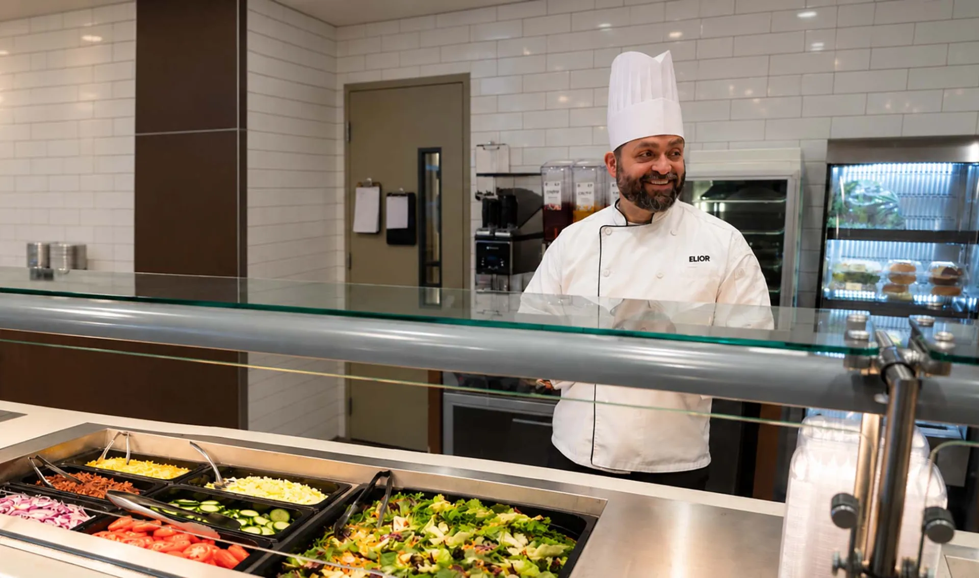 A smiling chef in a white uniform and tall hat standing behind a salad bar counter filled with various fresh vegetables and toppings in a cafe setting.