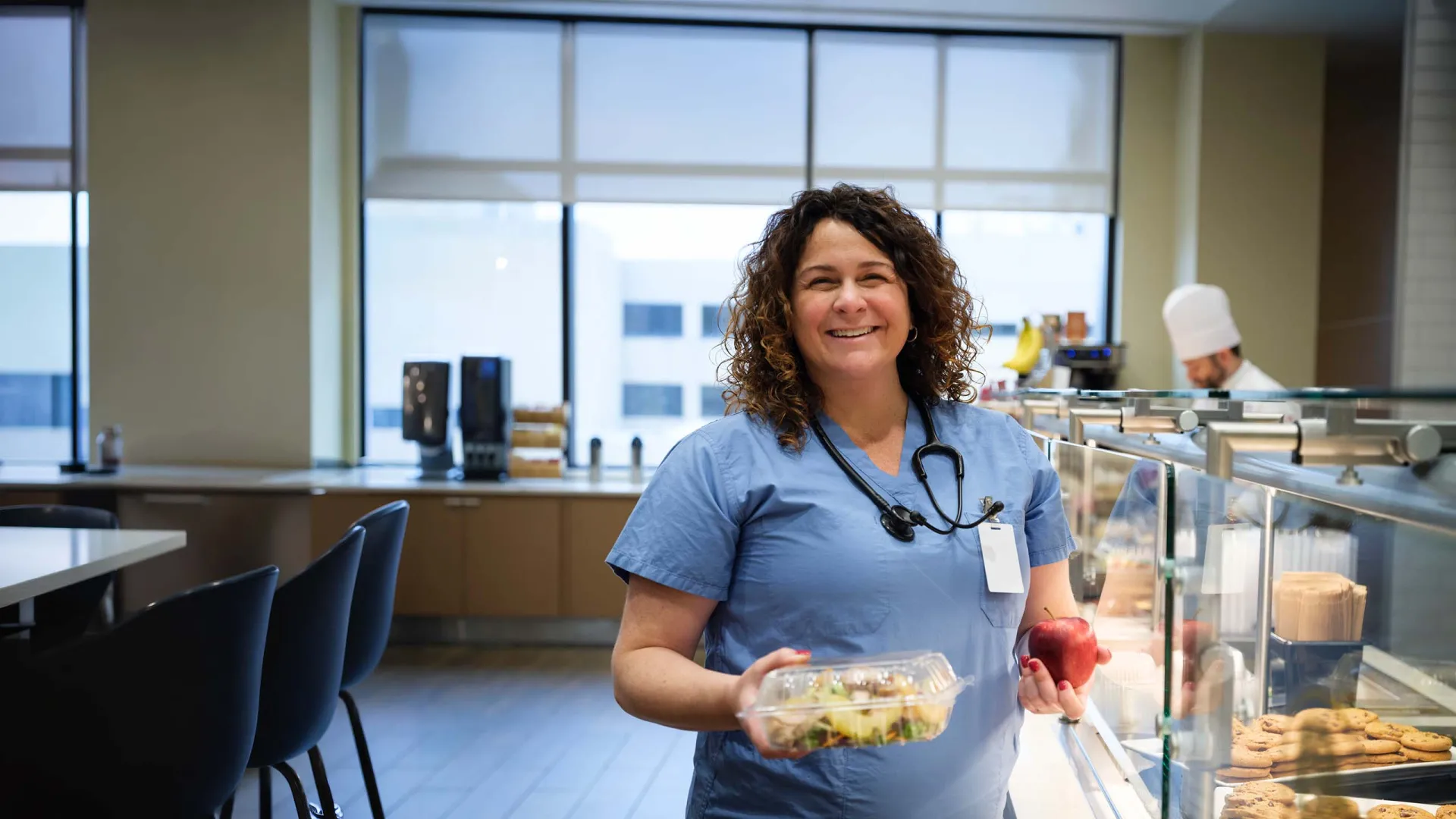 A smiling woman in blue medical scrubs holds a salad container and a red apple while standing at a cafeteria food counter.