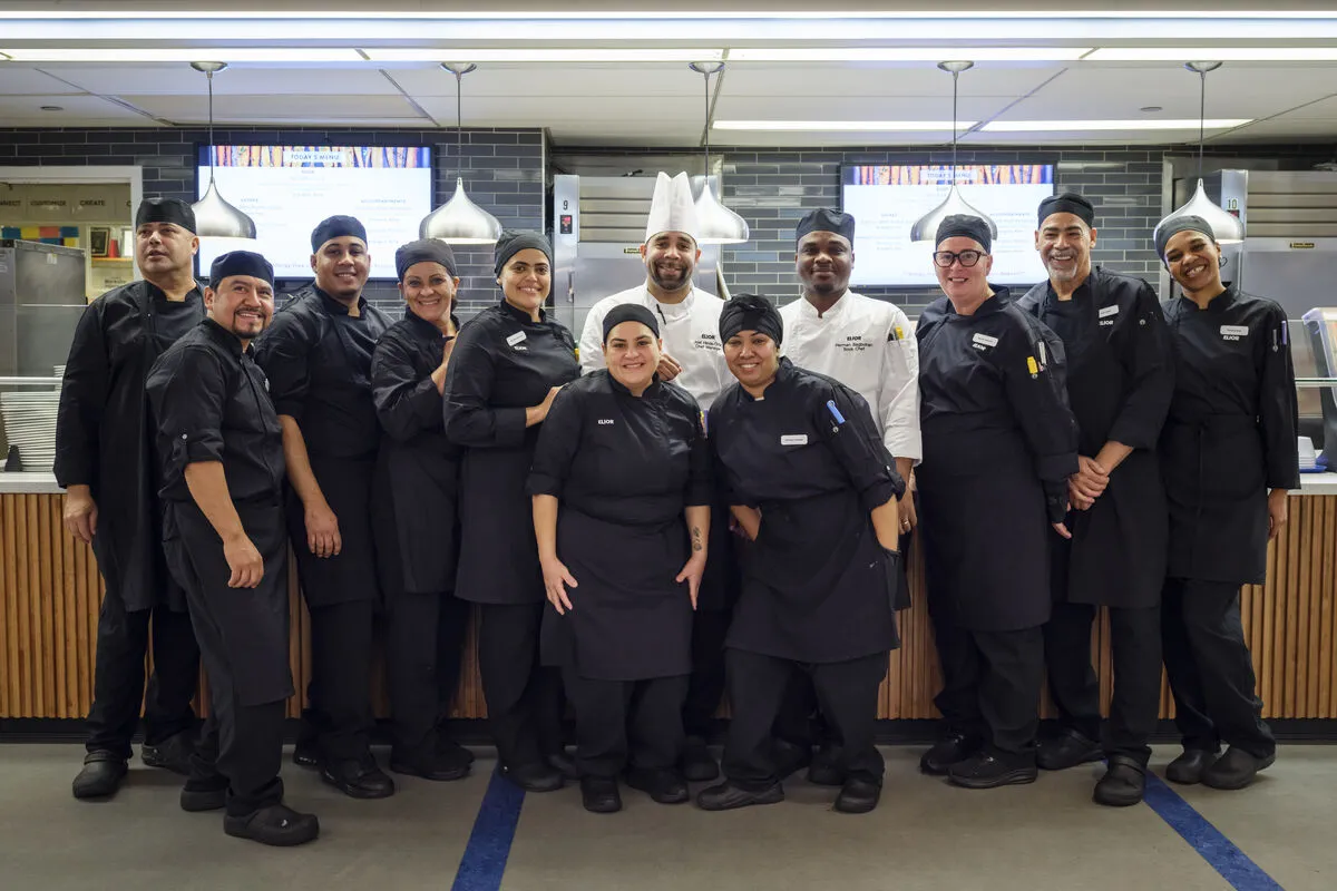 A diverse group of professional chefs and kitchen staff, dressed in black uniforms and white chef hats, are standing together and smiling in a modern commercial kitchen.