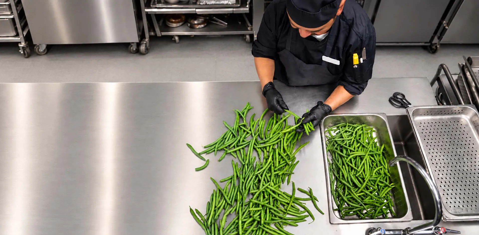 A chef wearing black gloves is sorting and preparing fresh green beans on a stainless steel countertop in a commercial kitchen.