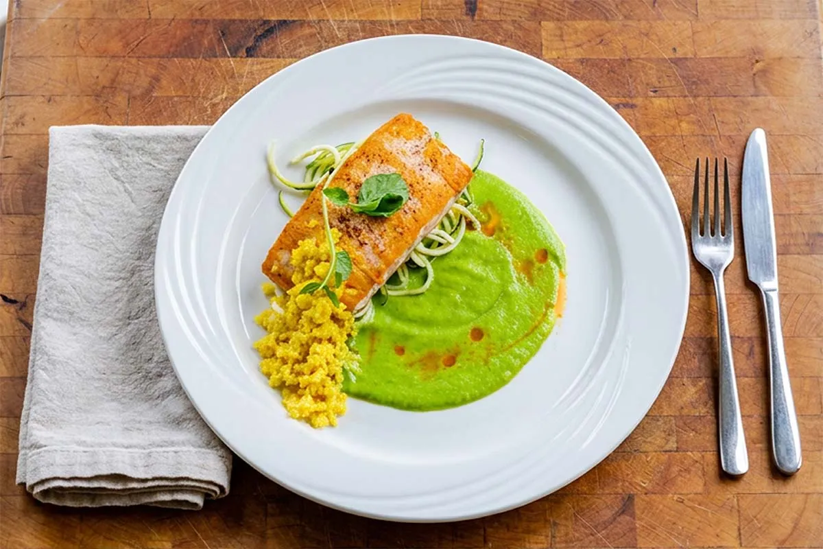 Overhead view of a plated salmon entrée with green vegetable purée, zucchini noodles, and grains on a rustic wooden table.