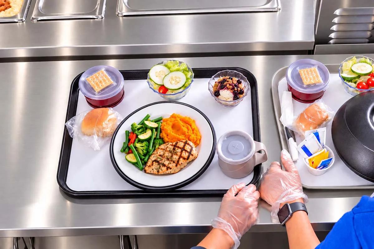 Overhead view of a hospital meal tray with grilled chicken, vegetables, mashed sweet potatoes, salad, bread, and beverages being prepared on a stainless-steel counter.
