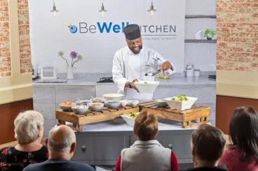 Chef demonstrating a cooking presentation to an audience for a BeWell Kitchen event.