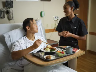 A patient in a hospital bed is eating a meal while smiling and talking with a healthcare worker standing beside the bed.