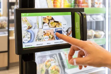 Hand using a touchscreen kiosk to browse food options in a self-service market.