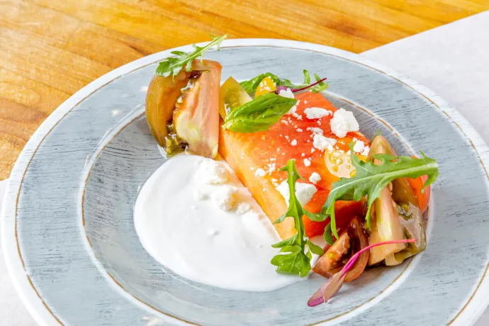 A plated dish consisting of a slice of watermelon topped with crumbled cheese and greens, accompanied by tomato wedges and a dollop of creamy sauce, all served on a light-colored plate placed on a wooden surface.