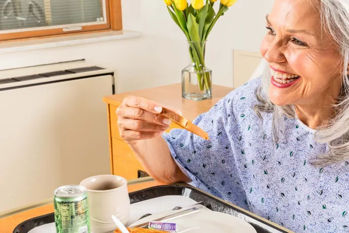A smiling hospital patient enjoys a meal while seated beside a tray in her room.