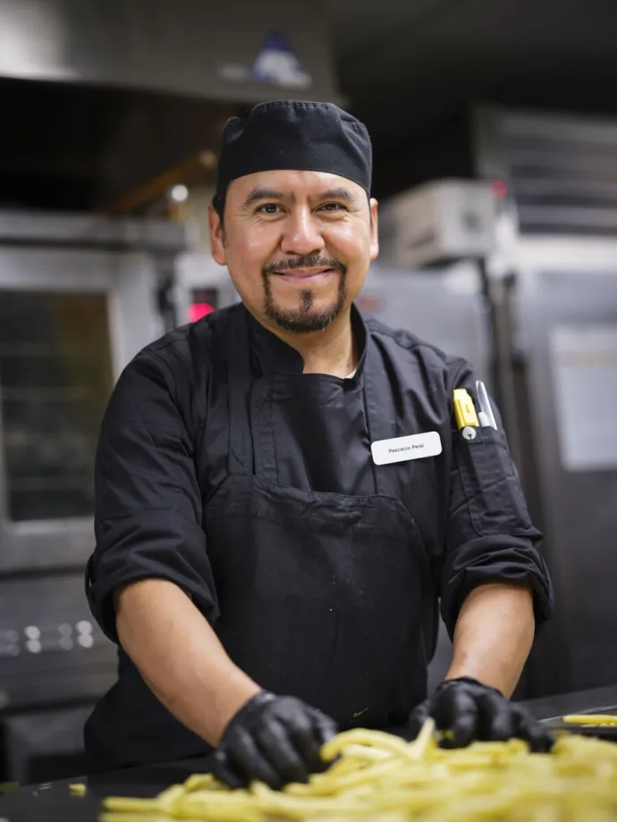 The image shows a chef wearing black attire and gloves, smiling while preparing food in a commercial kitchen.