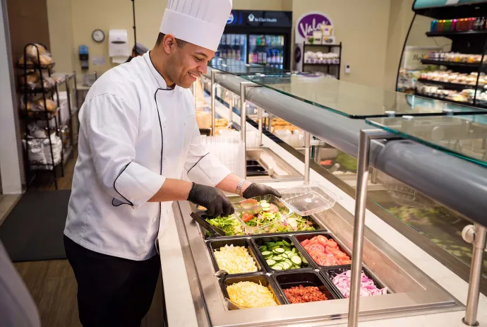 A chef wearing a white uniform and hat is preparing a fresh salad behind a glass counter in a cafeteria.
