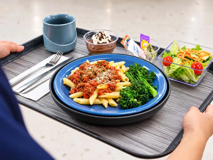 A person is holding a tray with a meal consisting of pasta with meat sauce, steamed broccoli, a small salad, a dessert, and a drink cup.