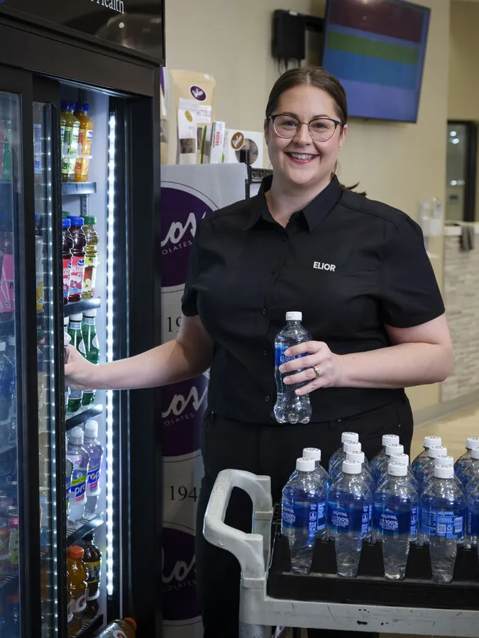 A woman wearing a black shirt is smiling while holding a water bottle and standing next to an open refrigerated display case filled with drinks in a well-lit cafe setting.