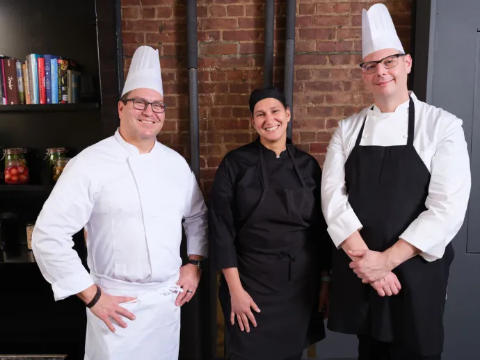 Three chefs standing and smiling in a kitchen setting with exposed brick walls and shelves holding jars and books.