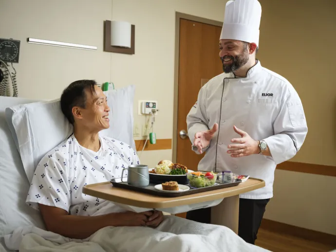 A chef in a white uniform and hat is smiling and talking to a male patient who is sitting up in a hospital bed with a meal tray in front of him.